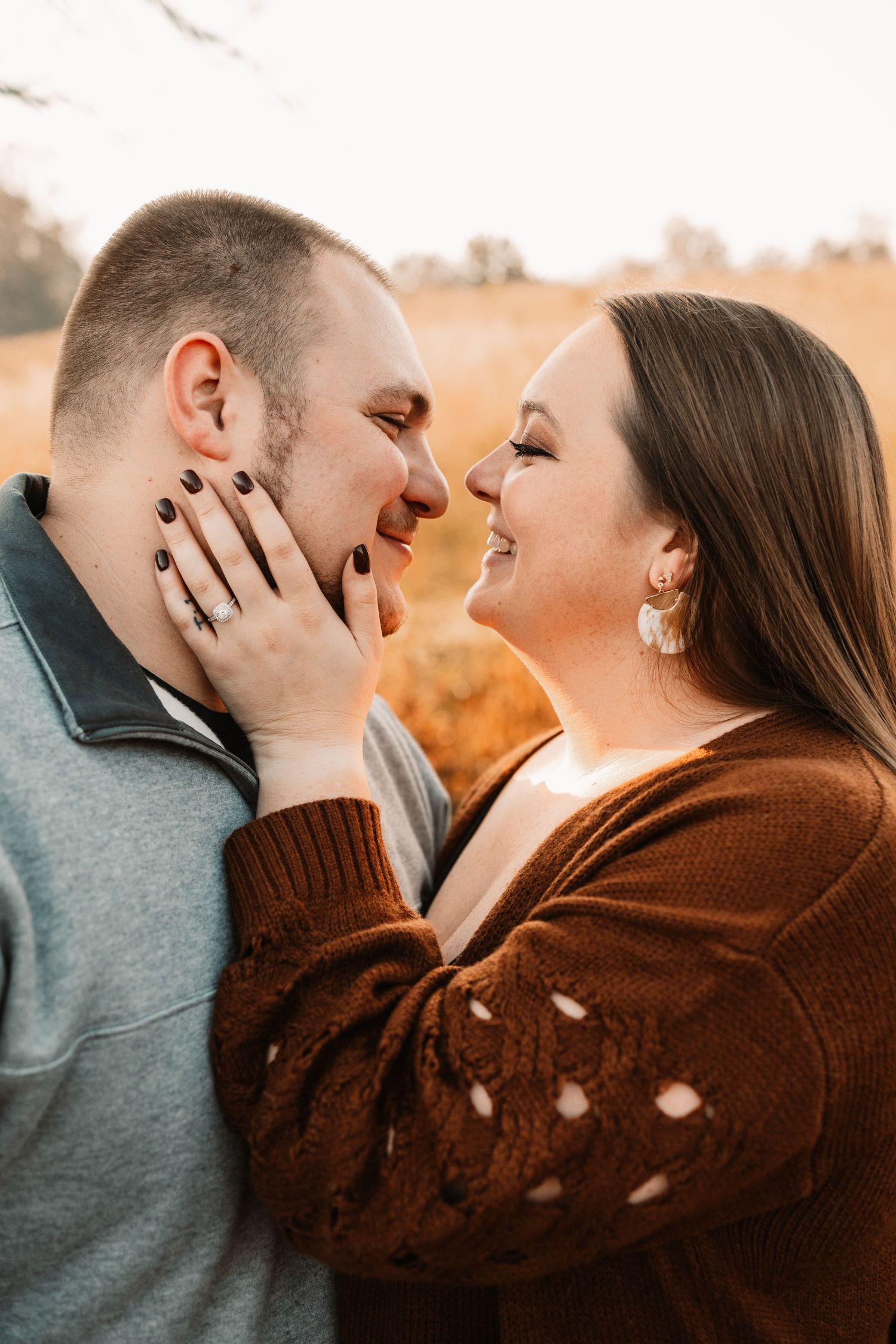 A romantic couple smiles at each other closely in a golden autumn field during warm sunset light.