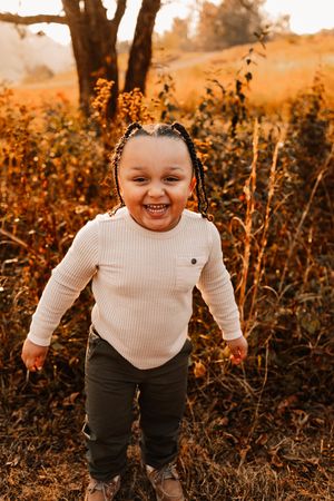 A happy young child with braids smiles broadly while standing in a warm autumn meadow at sunset.