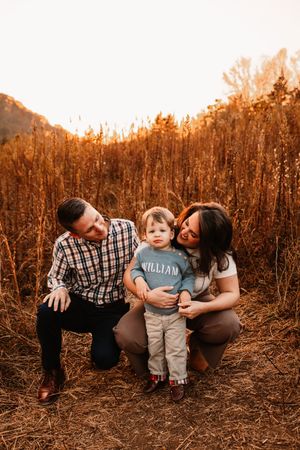 Family of three with toddler boy named William posing in tall golden autumn grass field.