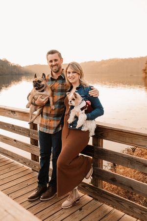 Smiling couple holding two dogs on wooden dock overlooking calm lake at golden sunset.