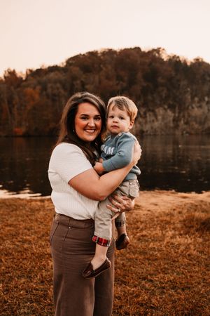 Smiling mother holding young toddler boy near autumn lake with colorful foliage background.