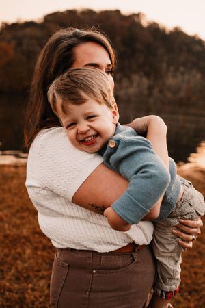 A joyful toddler boy laughs while being held by his brunette mother outdoors in a warm autumn setting.
