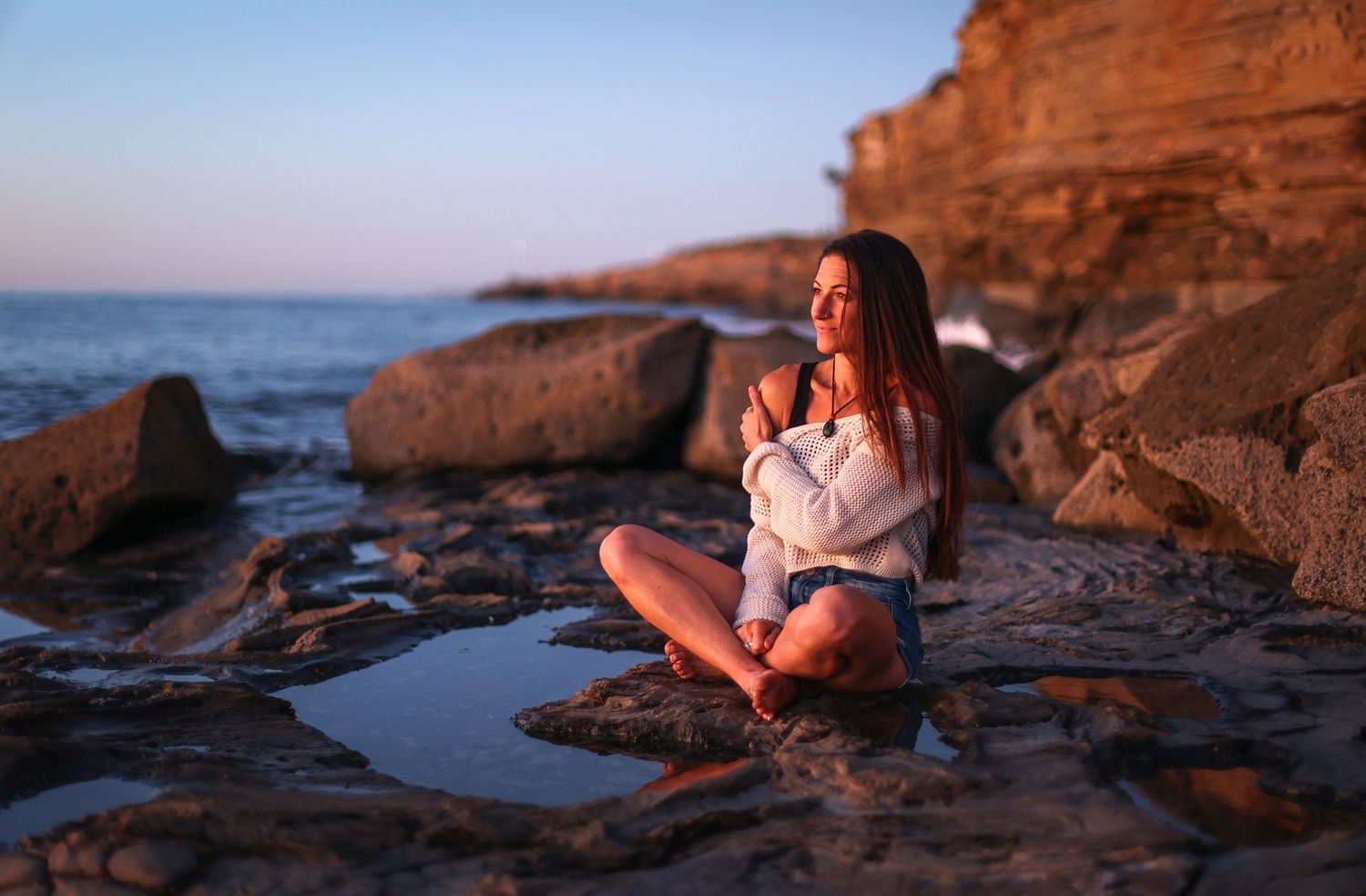 Woman meditating on rocky beach at sunset in white top and denim shorts.