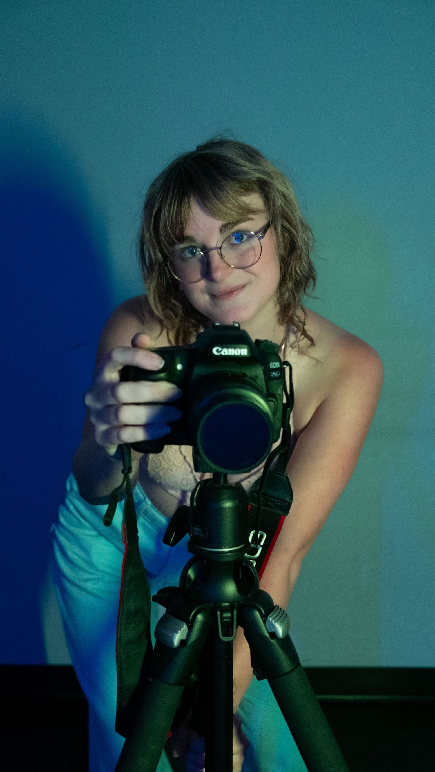 A photographer wearing glasses and blue top takes sequential self-portraits with a Canon DSLR camera in dim lighting.