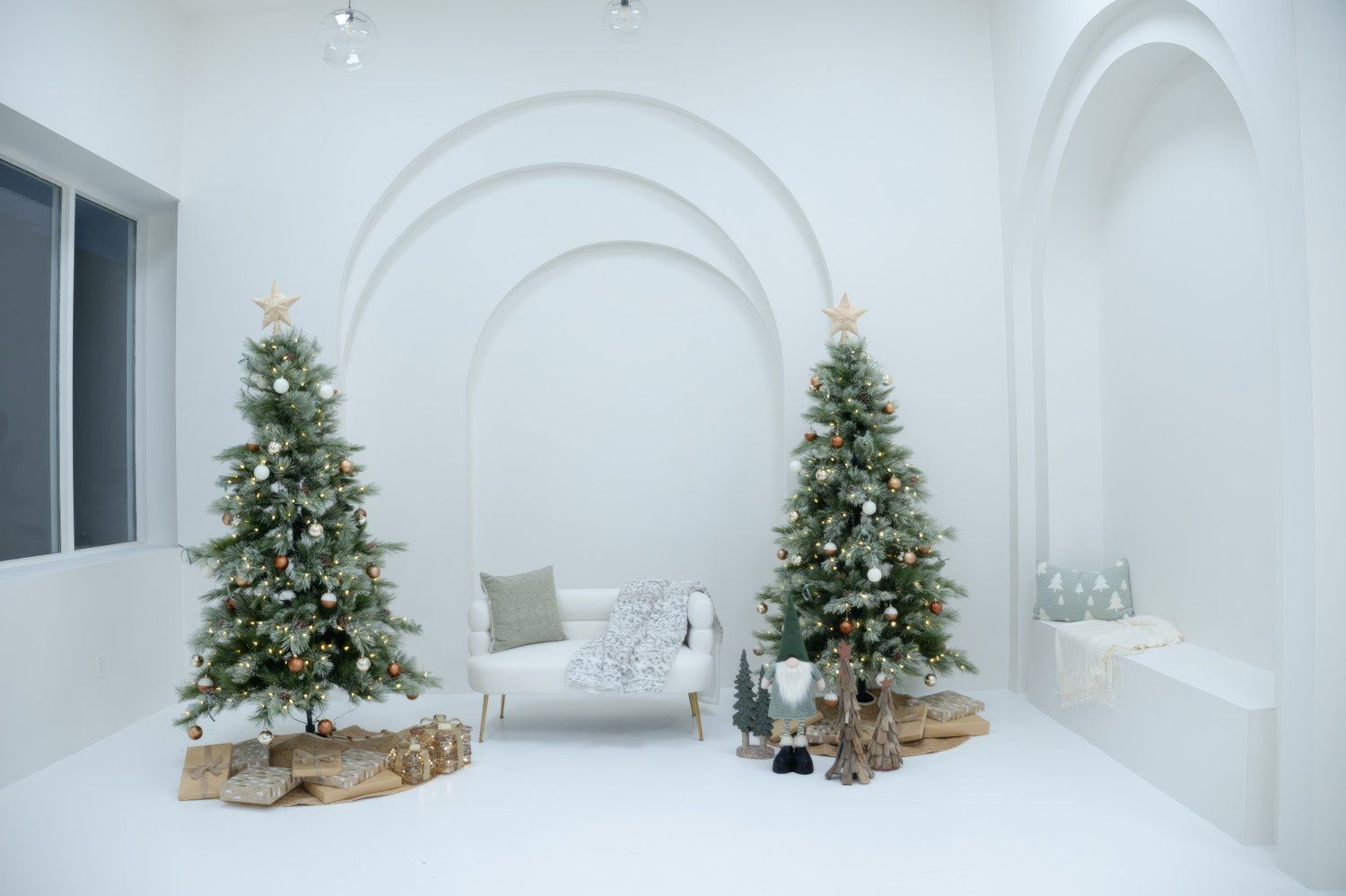 White minimalist room with two decorated Christmas trees flanking an elegant white chair with wrapped presents.