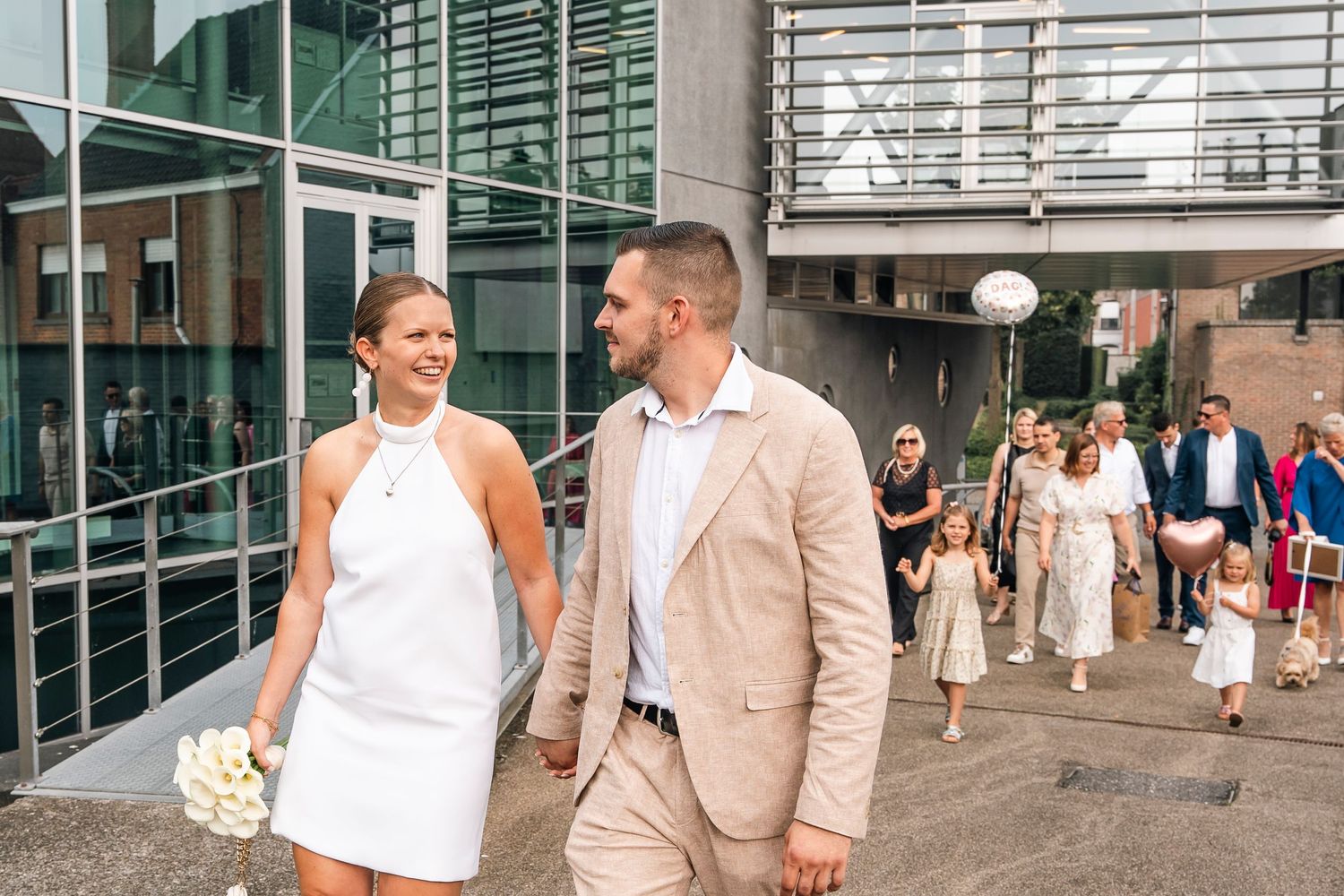 A wedding couple in white dress and beige suit walking outside a modern building entrance.