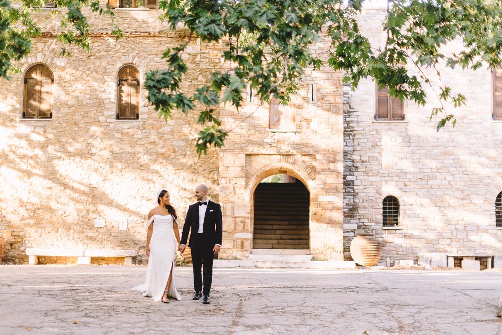 Bride and groom walk hand in hand outside a rustic stone building surrounded by lush greenery.