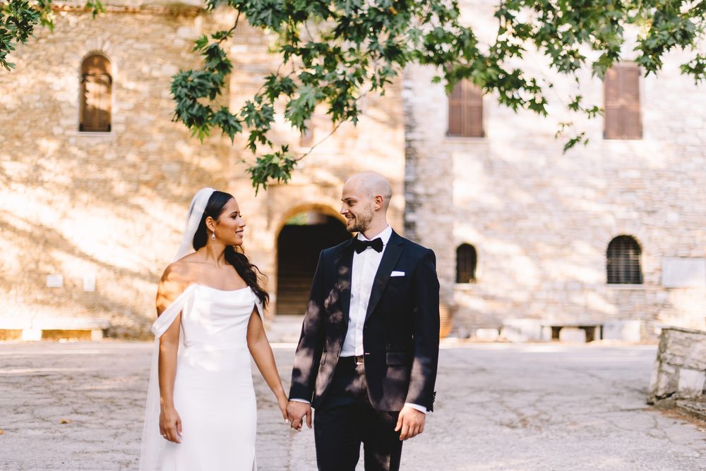 Bride and groom holding hands, smiling at each other in front of a rustic stone building with lush greenery.