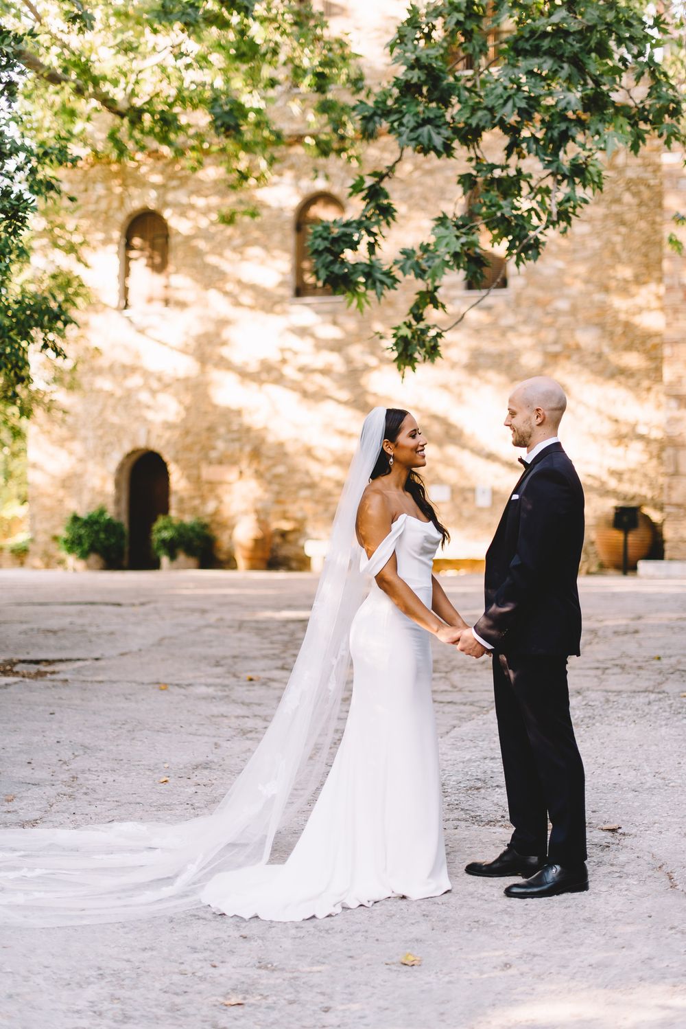 Bride in white off-shoulder gown with veil holds hands with groom in black tuxedo outside a stone castle.