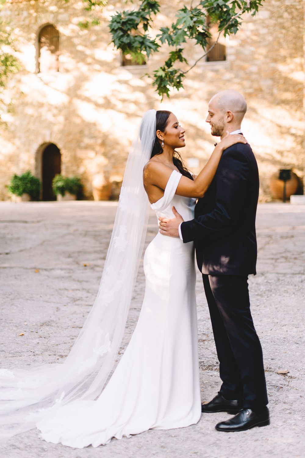 Bride and groom embrace in a sunlit courtyard, she wears a white off-shoulder gown with long veil.