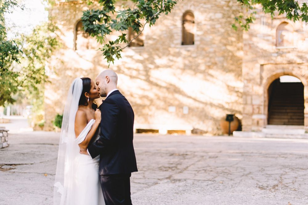 Bride and groom share a romantic kiss in front of a sunlit stone building, surrounded by lush green trees.