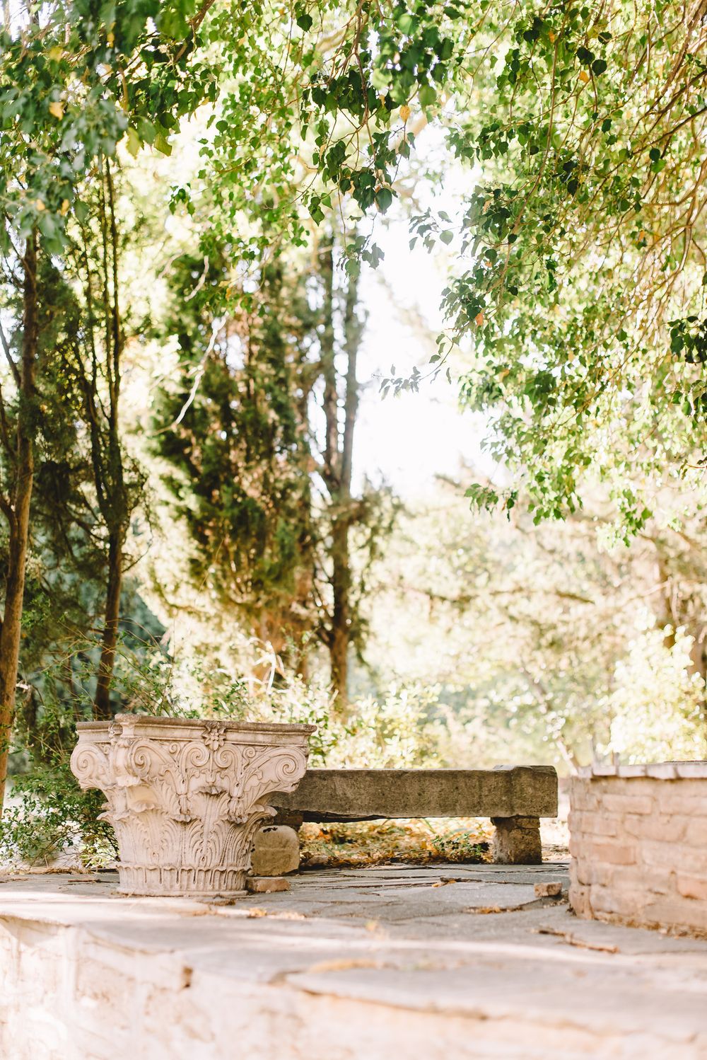 Ancient stone column capital and wooden bench surrounded by lush green trees in a sunlit garden.