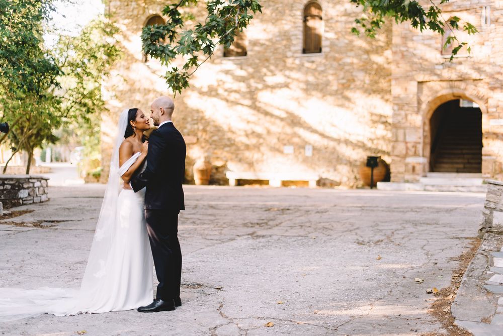 Bride and groom share a romantic kiss outside a sunlit stone castle surrounded by lush green trees.