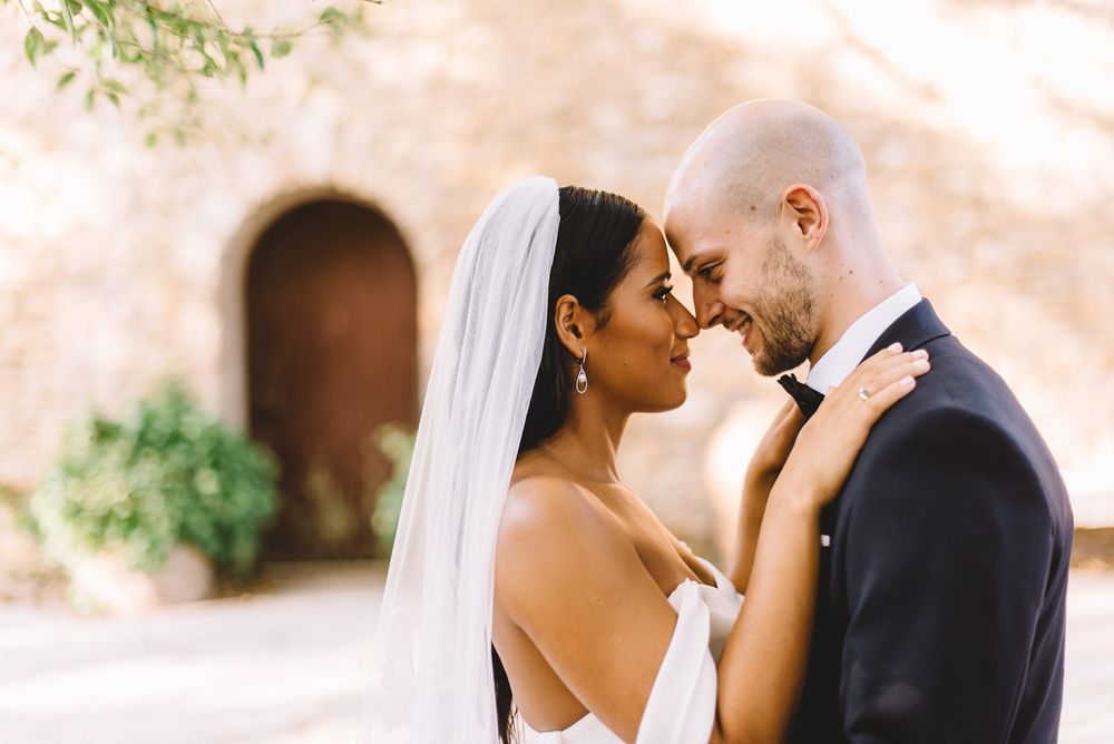 Bride and groom share an intimate moment, touching foreheads, with bride in white gown and veil outside a stone building.