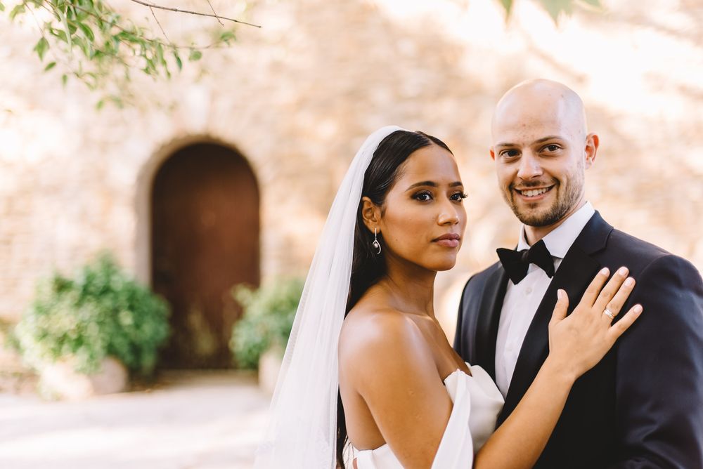 Bride and groom pose elegantly in wedding attire before a rustic stone archway, bride in white gown with veil.
