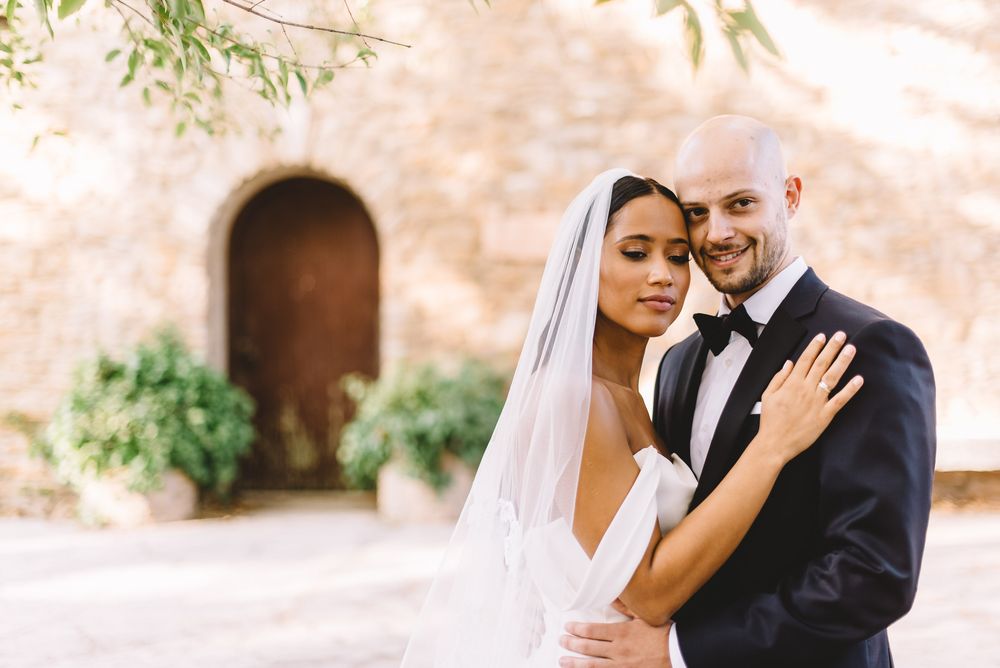 Bride in white gown and veil embraces groom in black tuxedo outdoors near stone building.