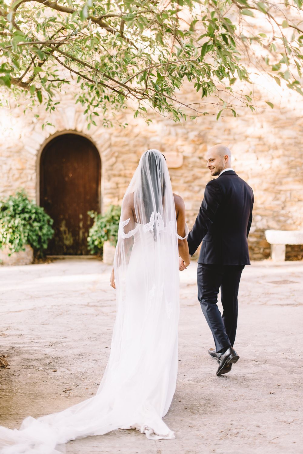 Bride and groom walking hand in hand toward a rustic stone building, groom smiling back at camera on their wedding day.