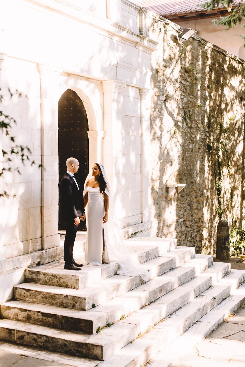 Elegant couple in formal attire standing on stone steps outside a historic white building with arched doorway.