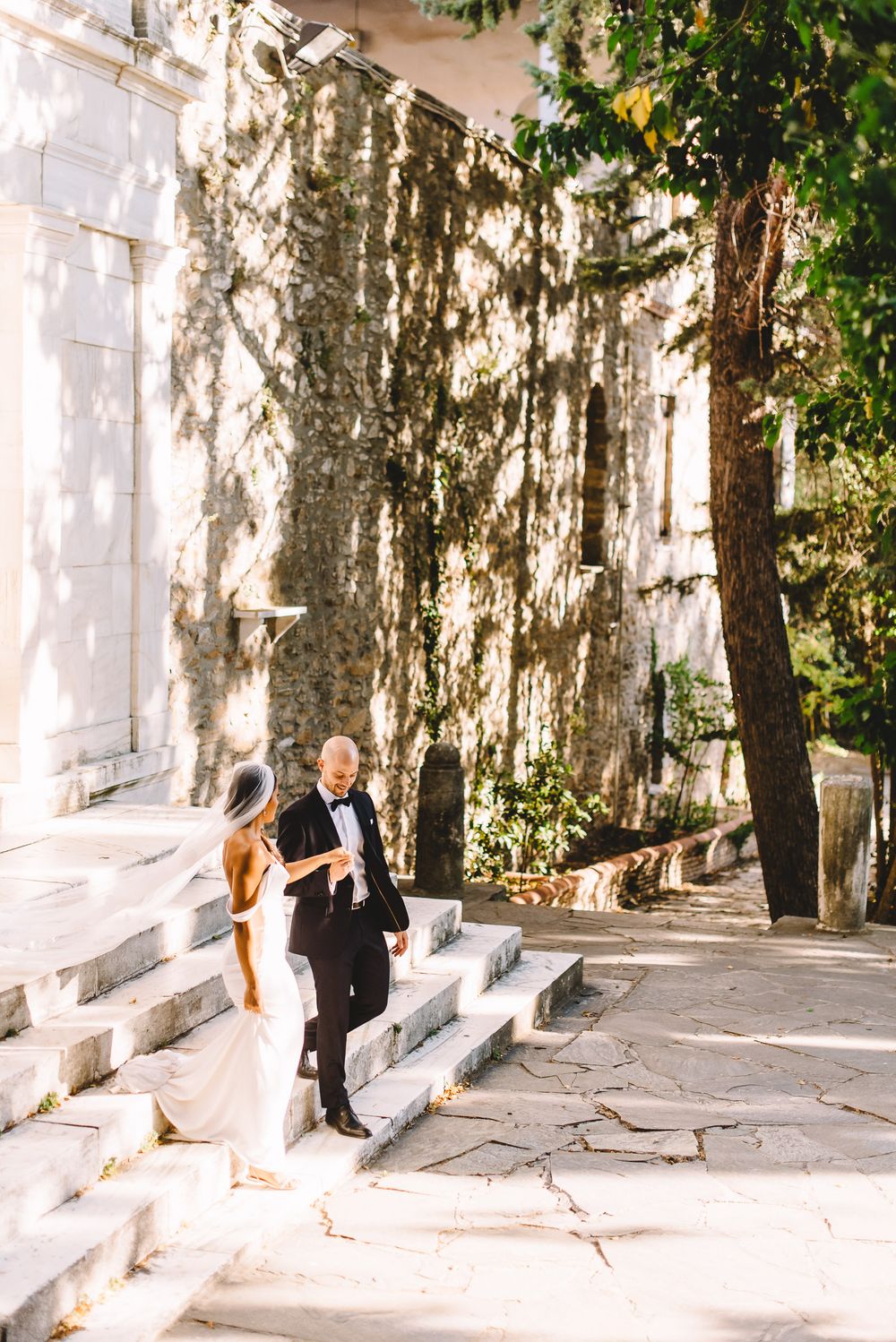 Bride and groom walking down stone steps near an ivy-covered wall in a sunlit outdoor wedding setting.
