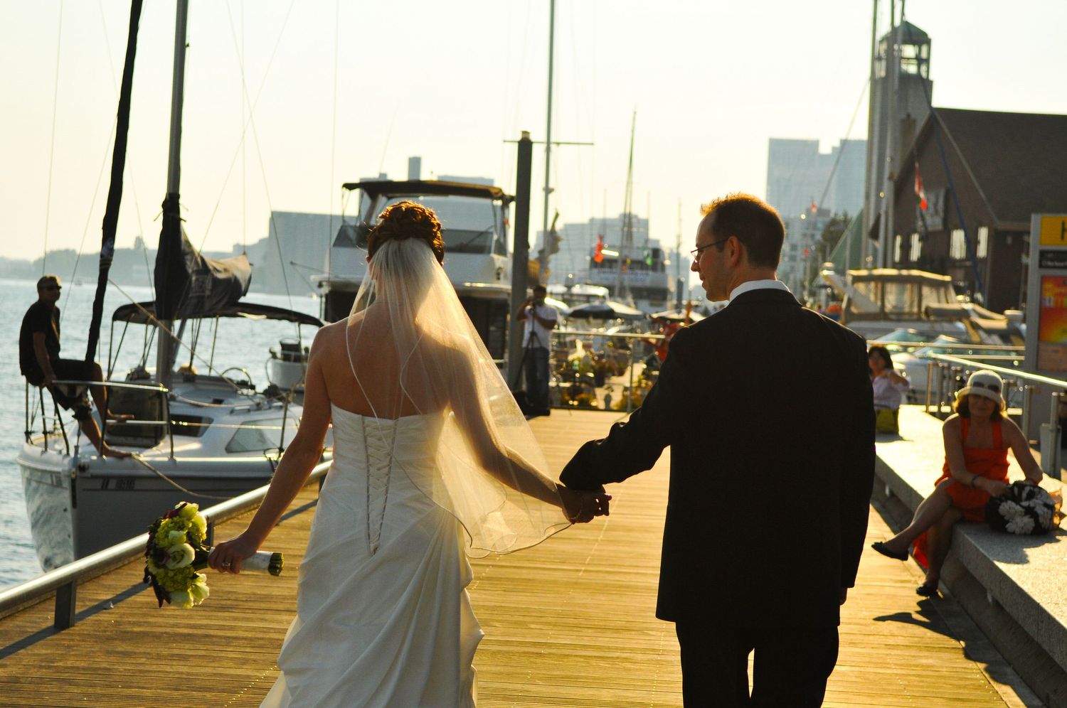 A bride and groom walking hand in hand on a marina dock at sunset with boats in the background.
