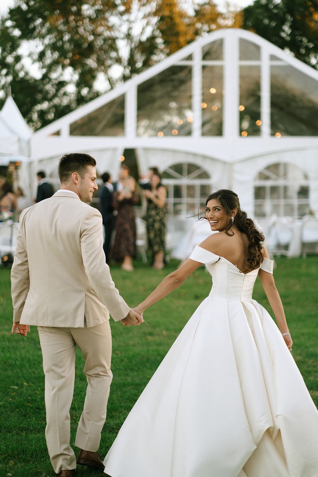 Bride in off-shoulder white ball gown holds hands with groom in beige suit walking toward elegant outdoor wedding tent.