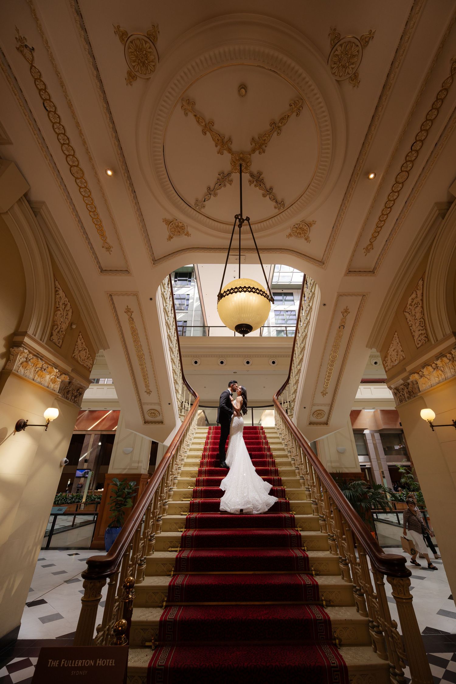 Bride in white gown and groom in suit standing before colonial sandstone building with vintage car.