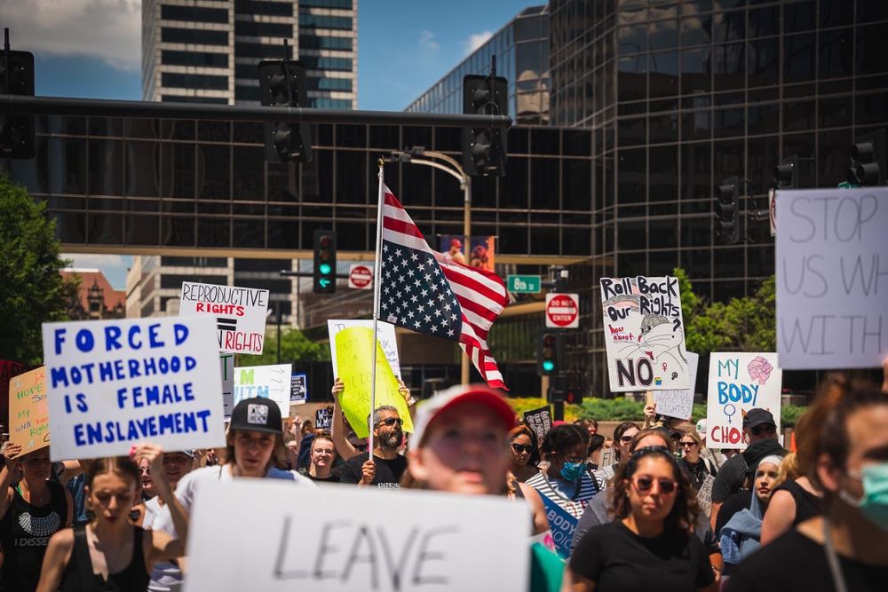 Crowd of protesters holding signs and American flag in front of glass building.
