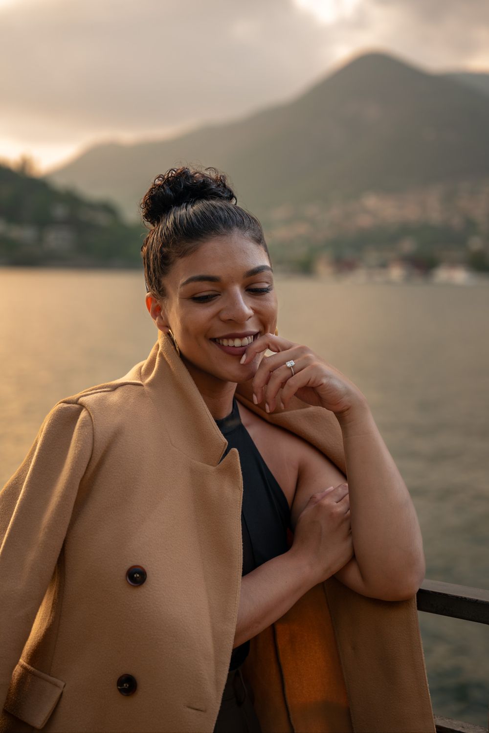 Candid sunset portrait by lake with mountains in background wearing camel coat.