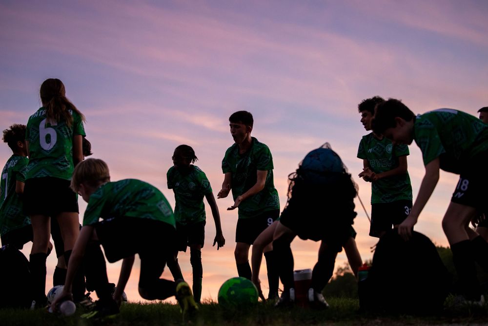 Silhouetted soccer team gathering at sunset with pink and purple sky.