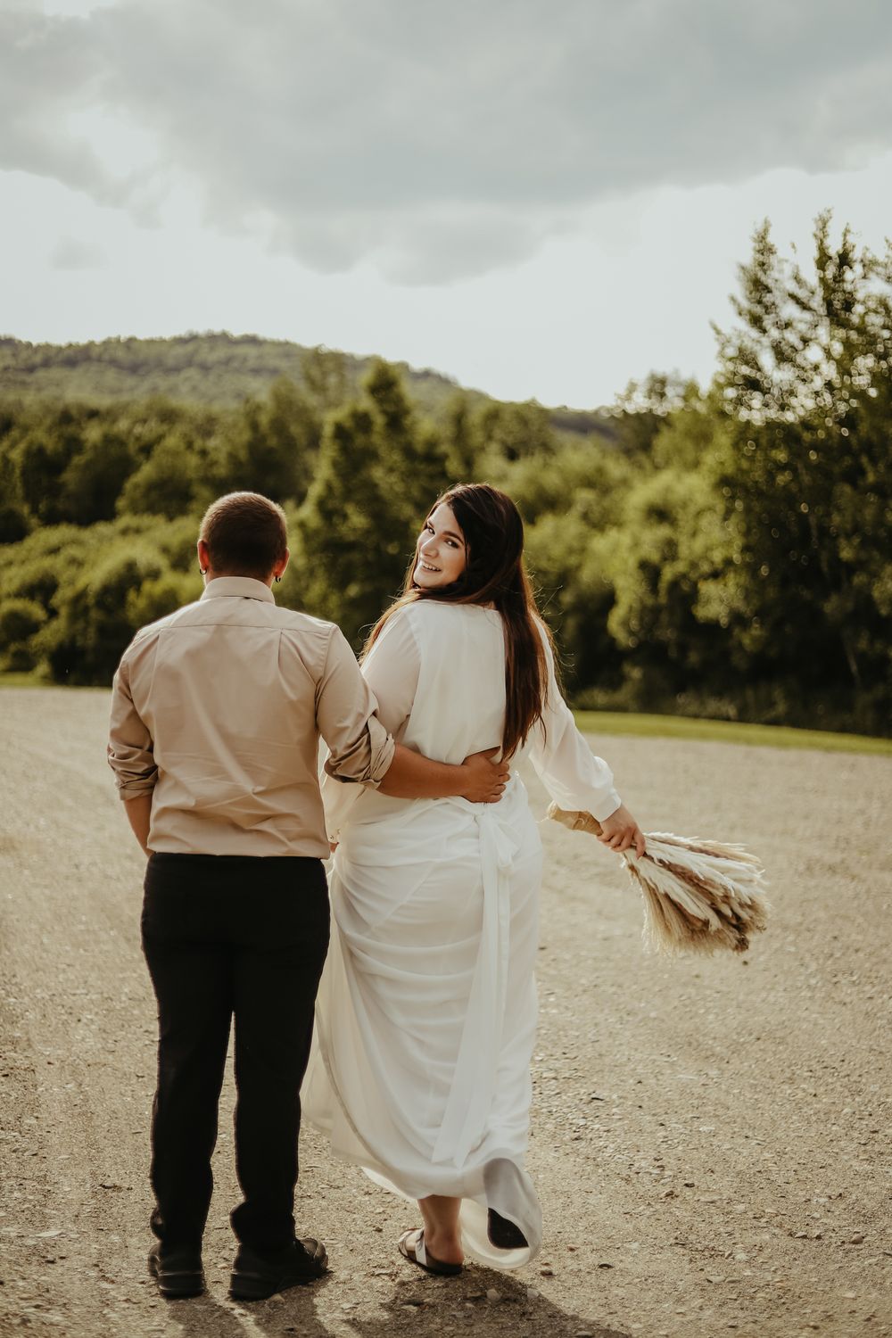 Couple walking on dirt path with dried flowers against mountain landscape.