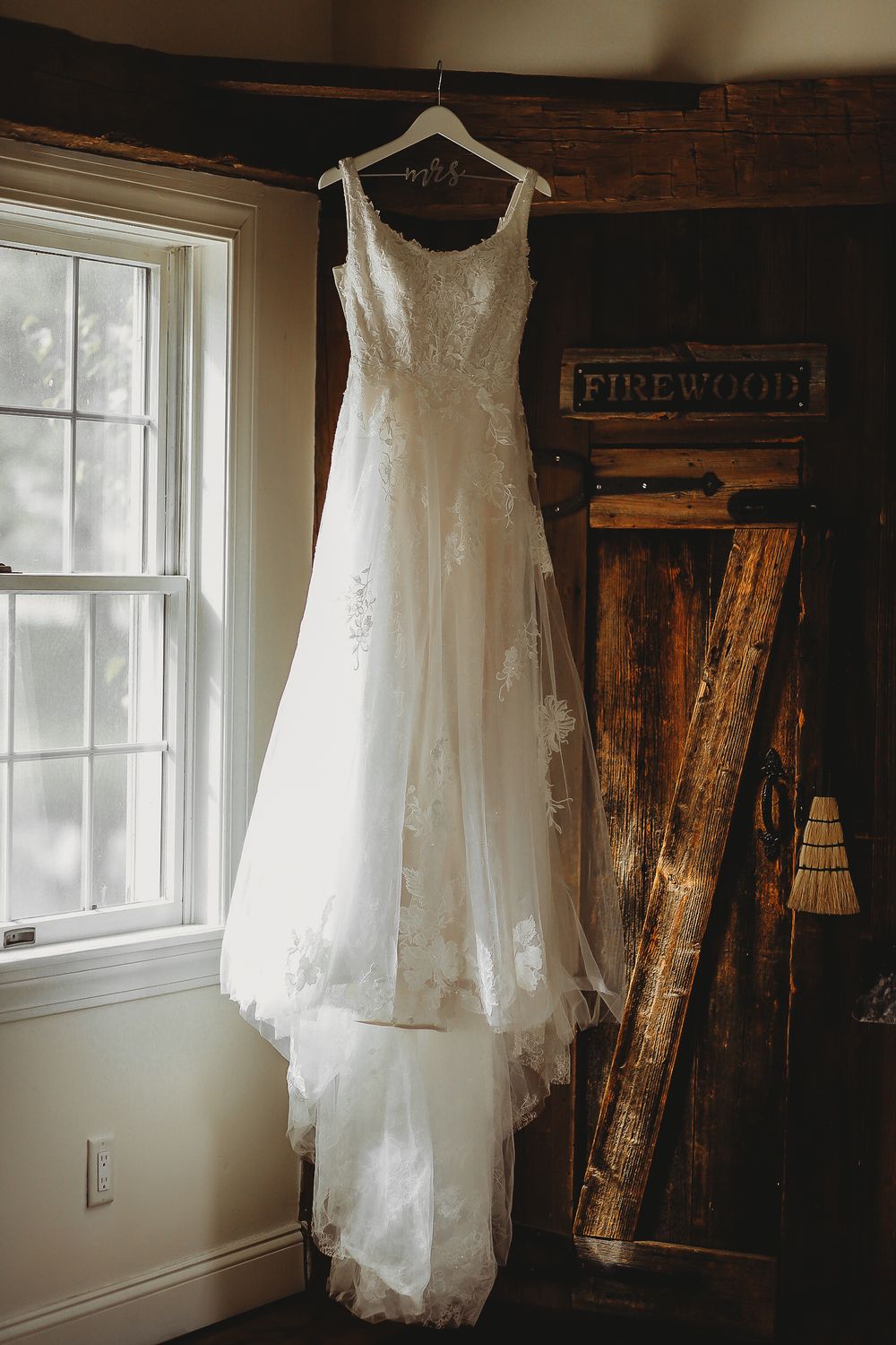An elegant white lace wedding dress hanging by a window in a rustic wooden room.