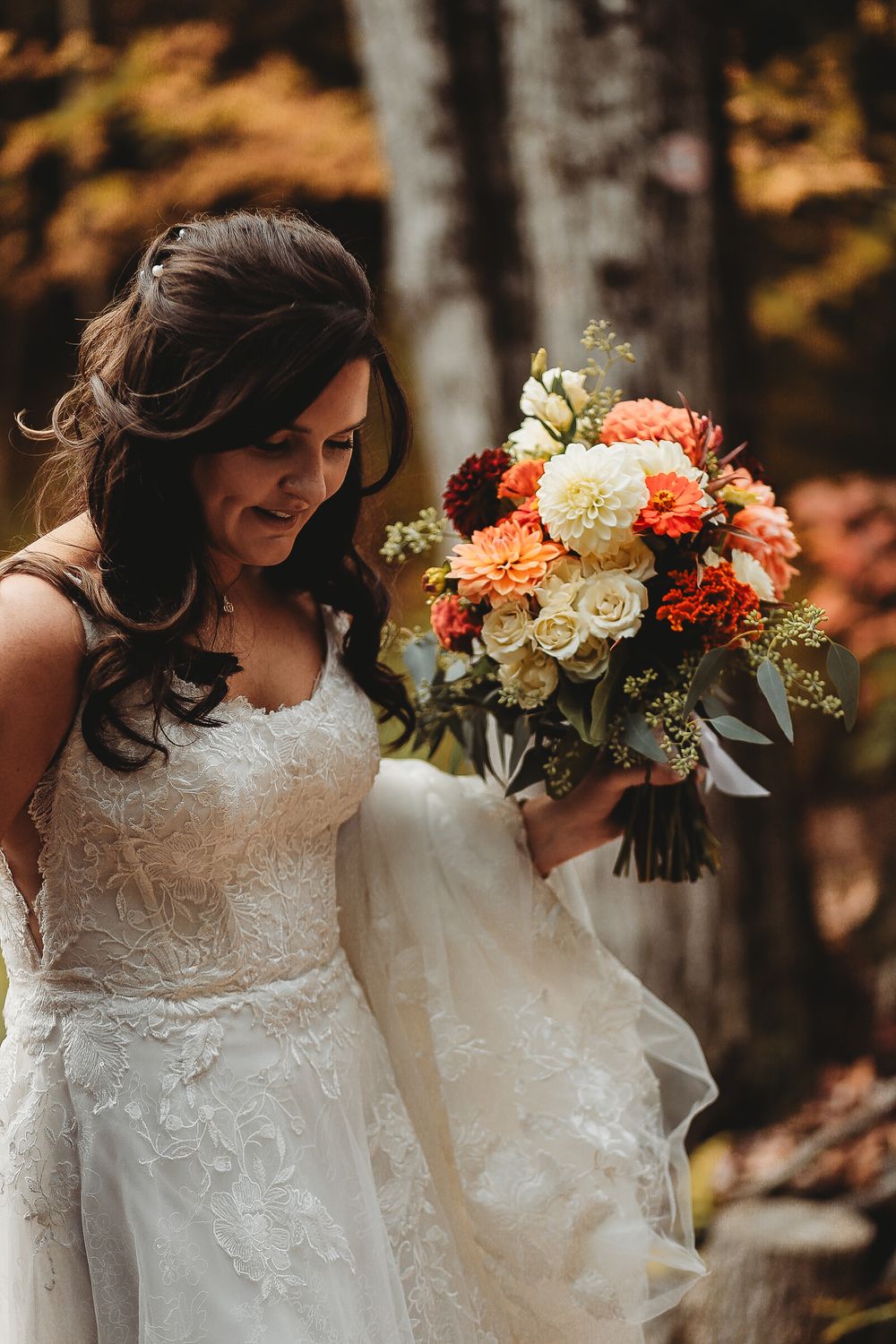 A bride in a lace gown holds a vibrant bouquet of coral and white flowers in an autumn setting.
