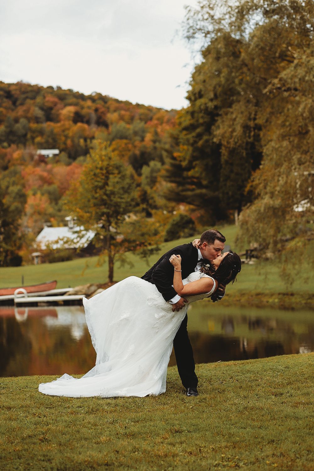 A romantic dip kiss between newlyweds on a grassy hill with autumn foliage and a lake in the background.
