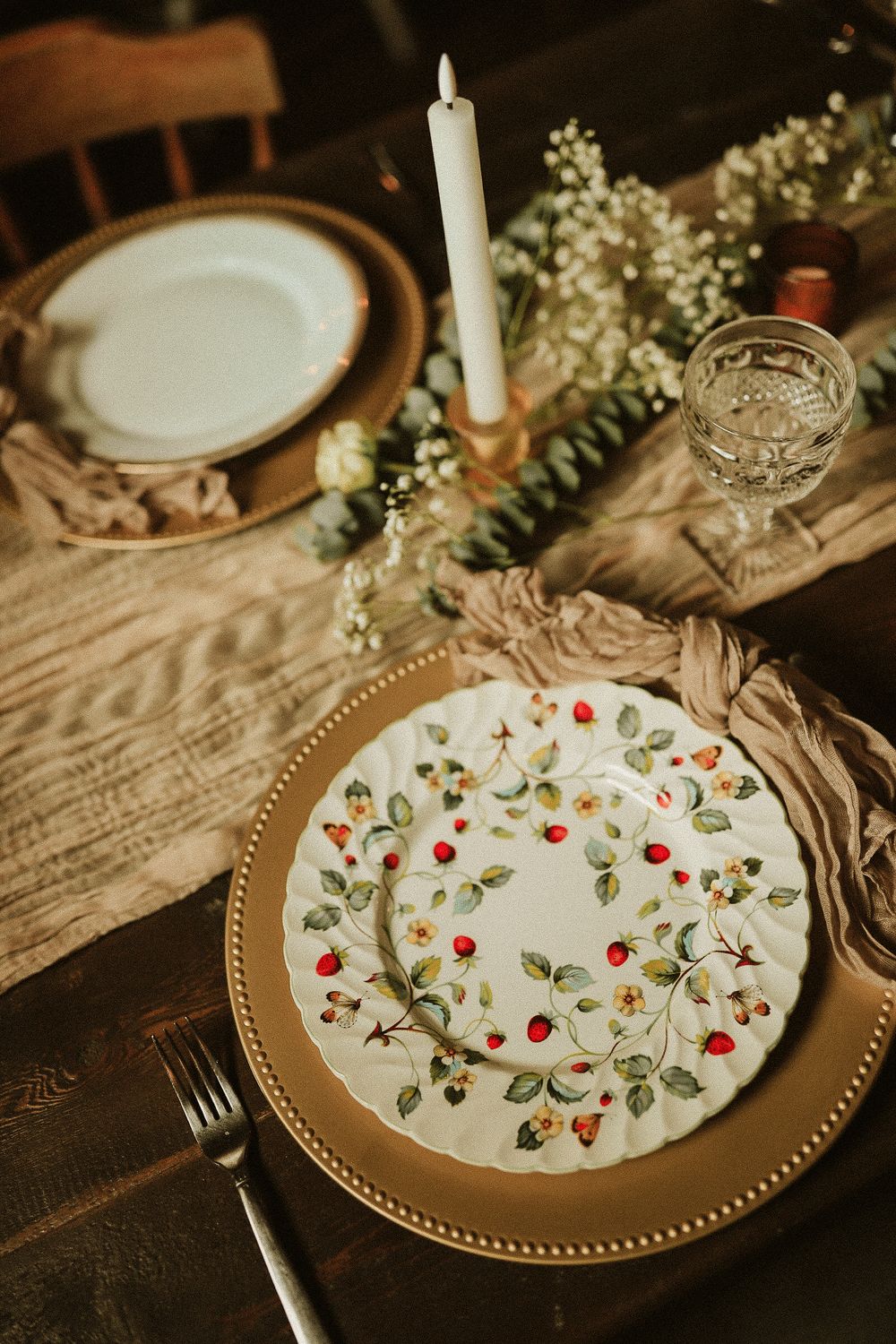 Romantic table setting with floral plate, gold charger, candle and delicate white flowers on wooden surface.