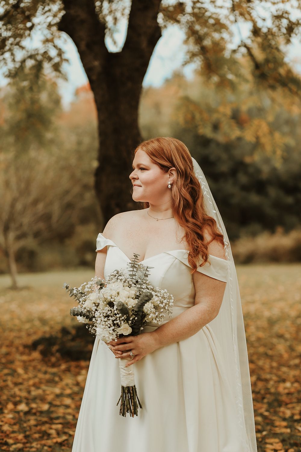 A bride in an off-shoulder gown holds a delicate bouquet against an autumn backdrop.