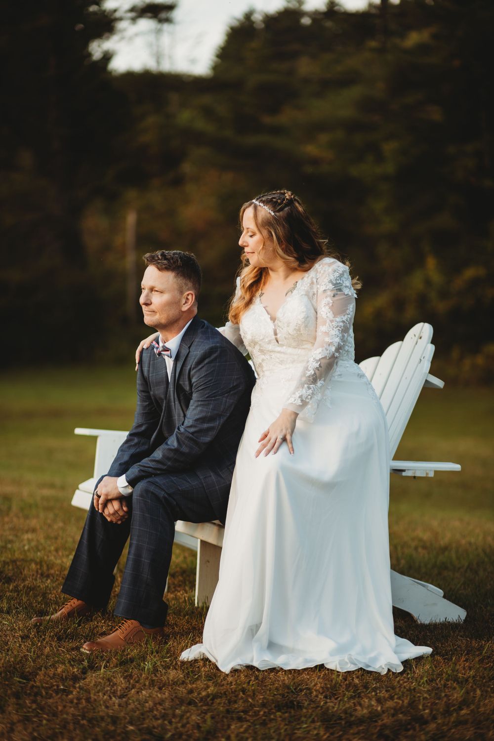 Wedding couple sitting on white Adirondack chair in outdoor setting.