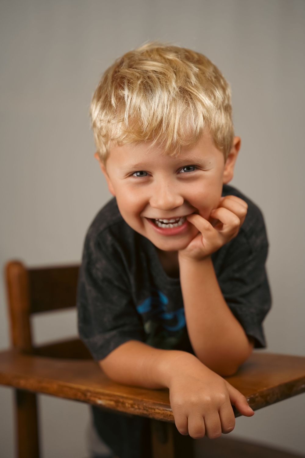 Young child in dark shirt leaning playfully on wooden table in casual indoor portrait series.