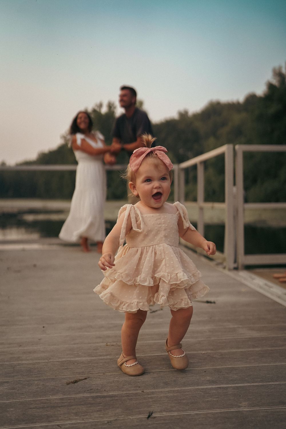 Baby in ruffled cream dress taking first steps on wooden deck.