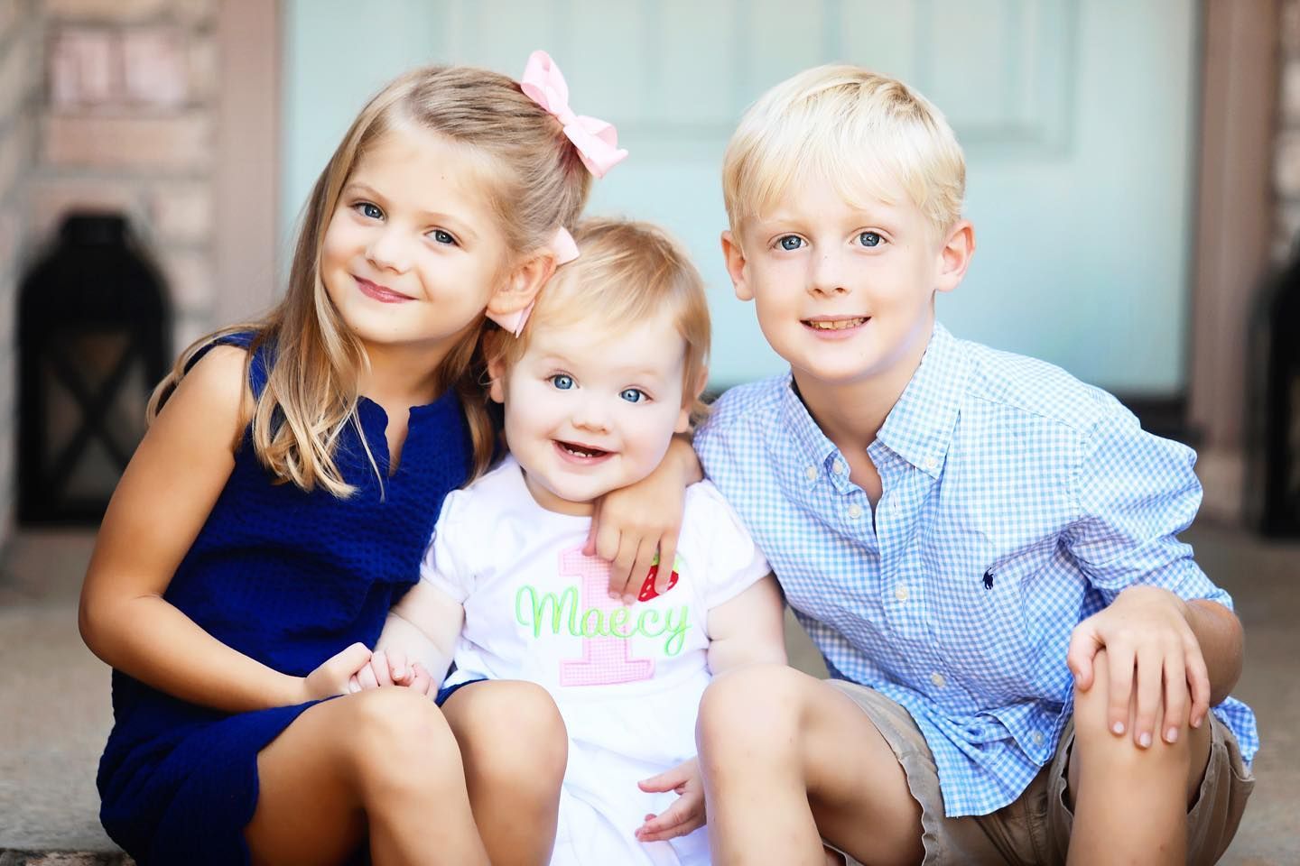 Three children sit together on outdoor steps wearing coordinated blue and white outfits.