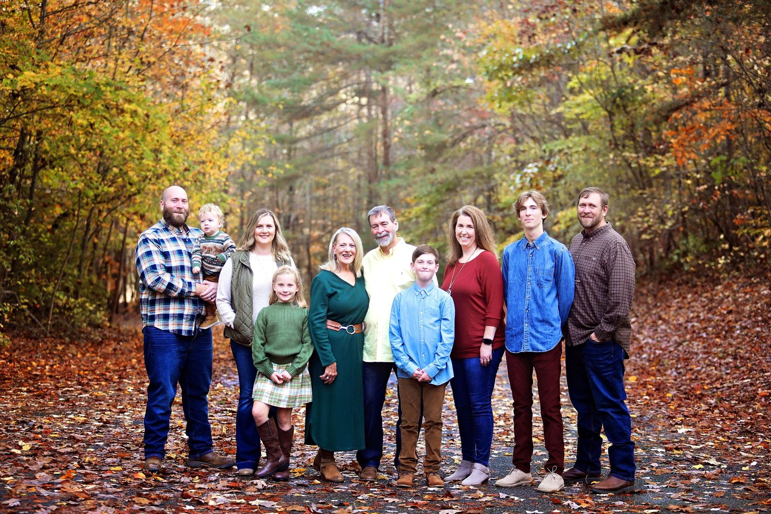 Large family group photo taken on fallen autumn leaves in a wooded setting.