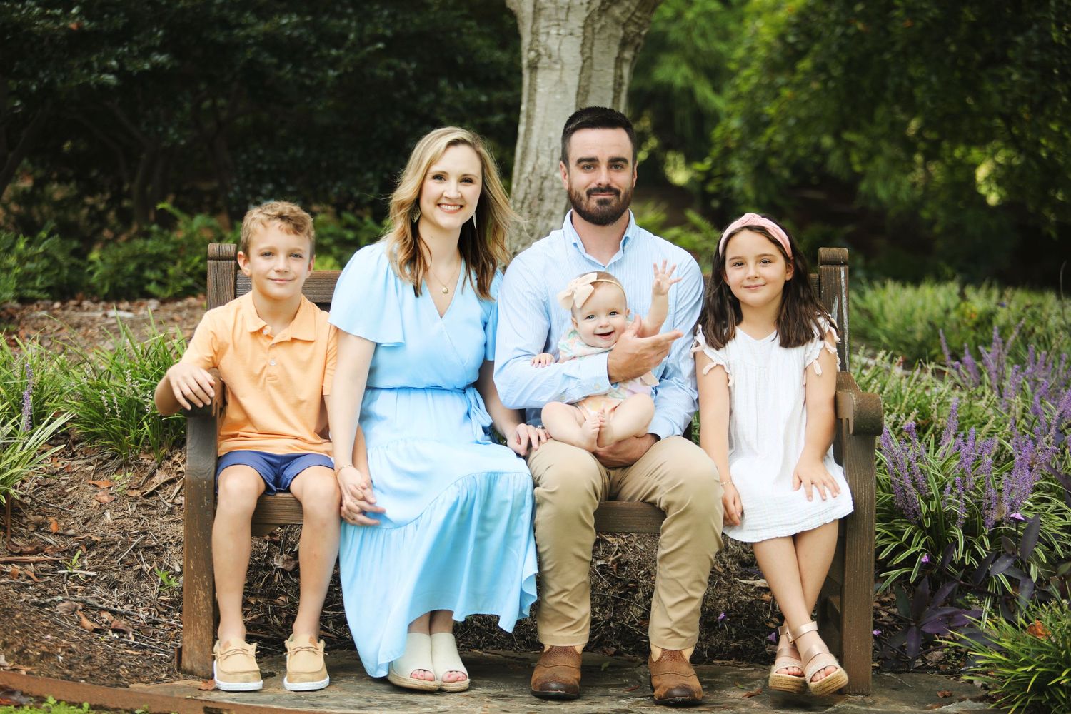 A family of five sits together on a wooden bench in a garden with blooming purple flowers.