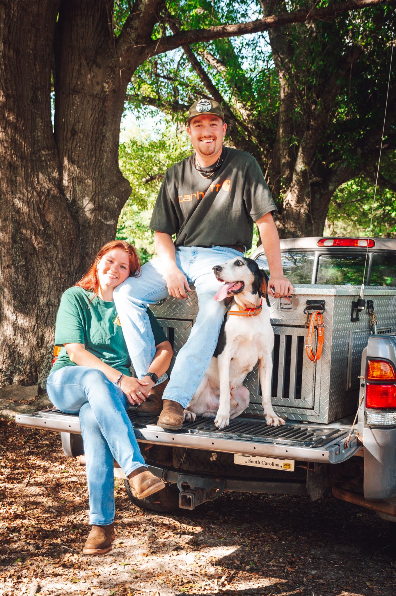 A couple relaxes on pickup truck tailgate with their spotted dog under shady trees.