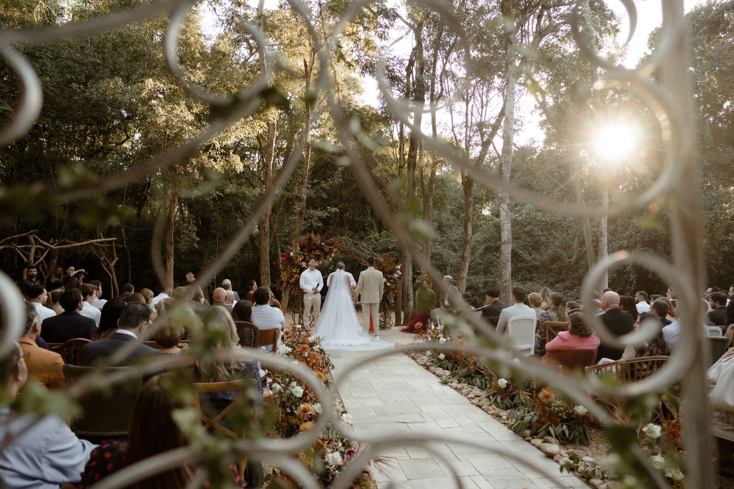 Wedding ceremony set in a sunlit wooded area with natural decorations.