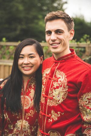 Couple wearing matching red and gold traditional Chinese wedding attire smiling outdoors.