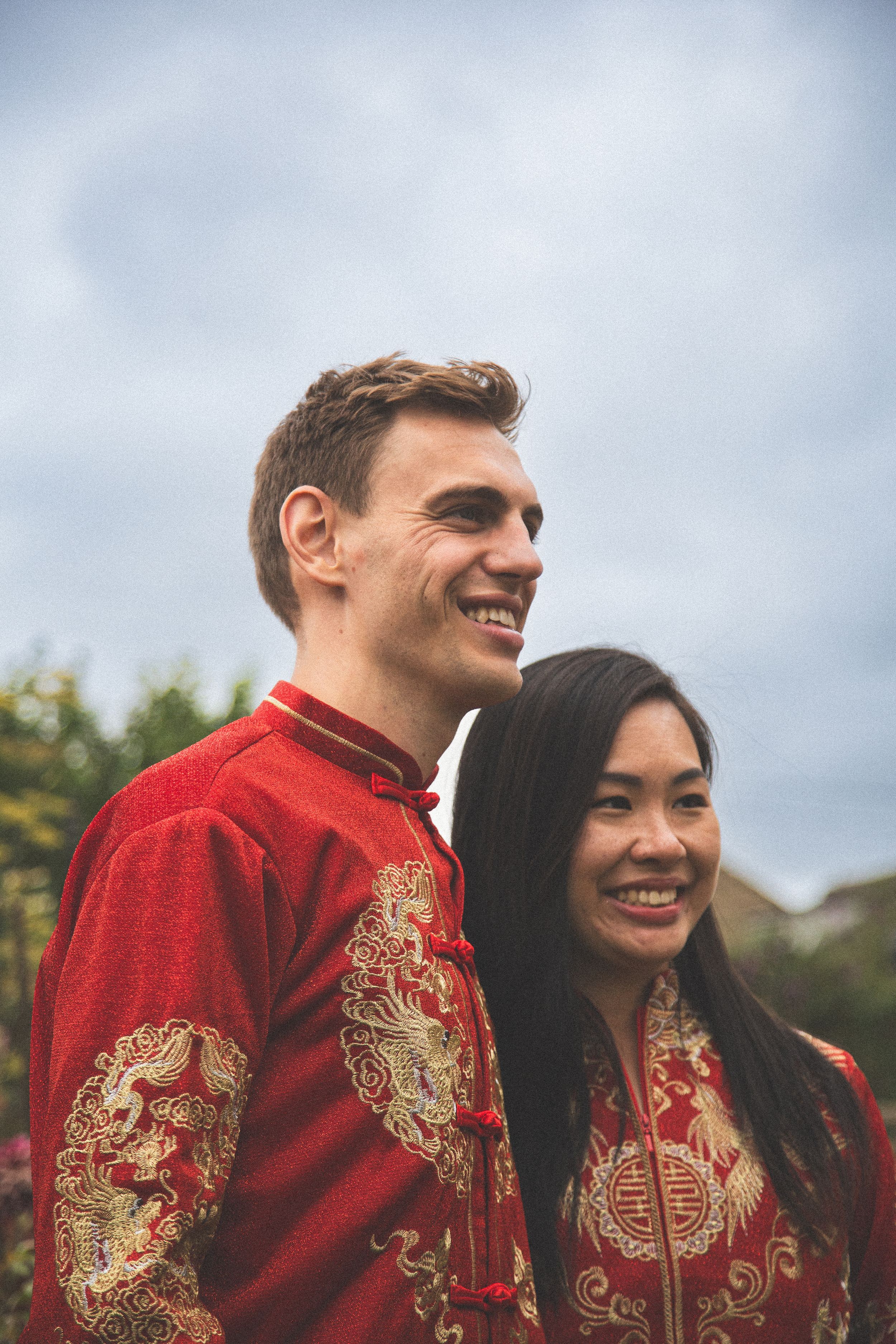 A couple wearing traditional red Chinese wedding attire with gold embroidery smiling outdoors.