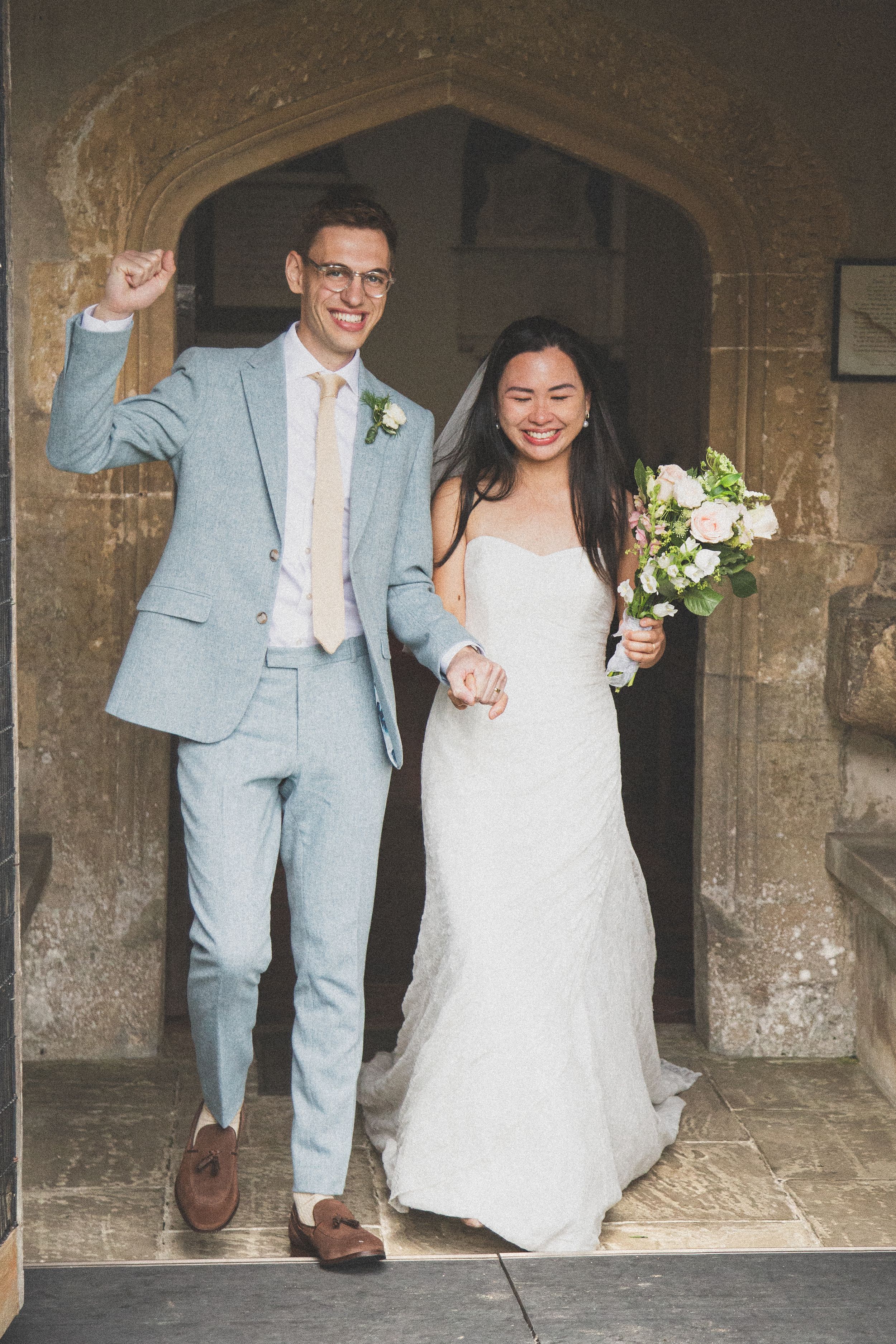 A bride in white and groom in light blue suit walking through stone archways.