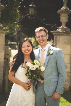 Happy newlyweds posing in a garden with stone pillars, bride holding a floral bouquet.
