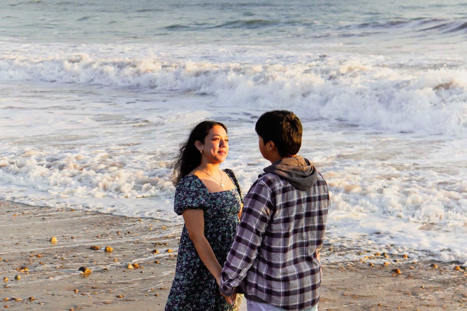 Couple holding hands while watching ocean waves crash on sandy beach.