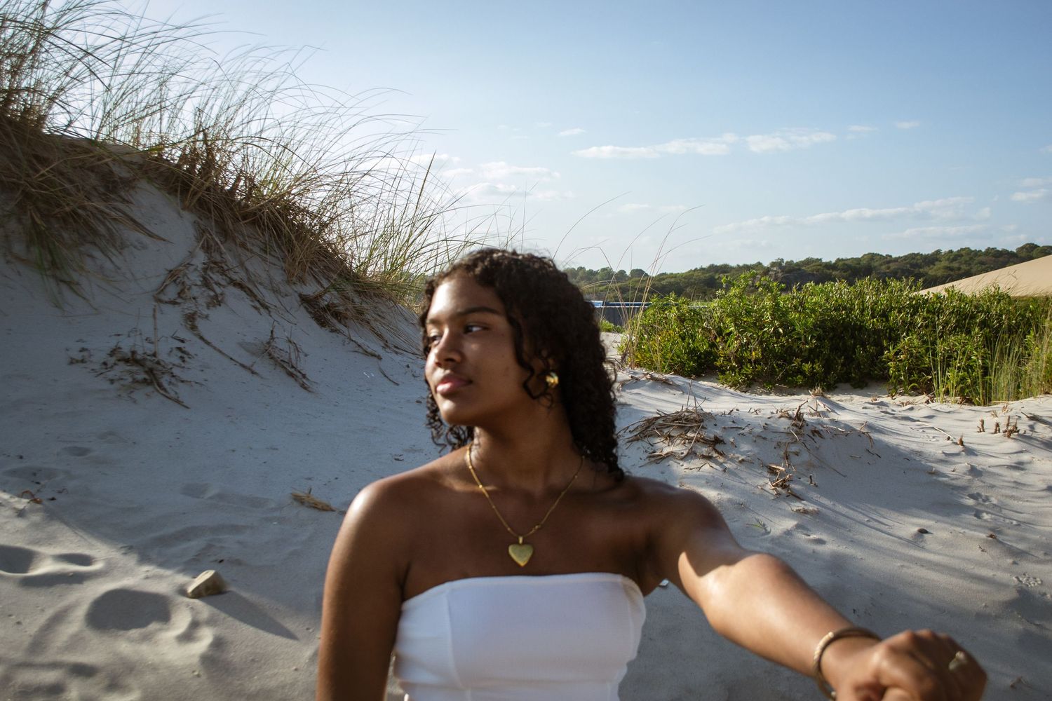 Person in white dress poses on sandy dunes with beach vegetation.