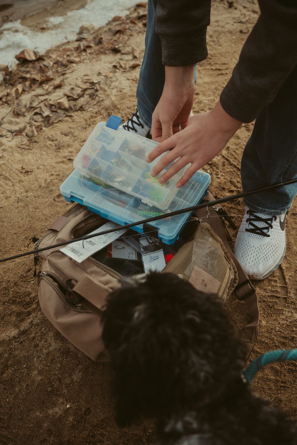 Hands organizing items in a blue container on dirt ground.